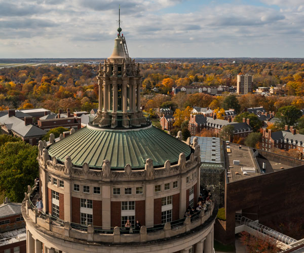 Aerial view of the University of Rochester's iconic Rush Rhees Library tower, featuring its domed roof and classical architecture. The surrounding landscape shows the campus buildings and autumn foliage extending into the distance under a partly cloudy sky.