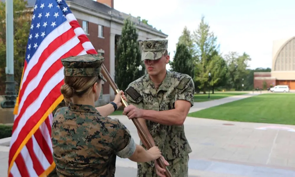 Female NROTC cadet in uniform hands off the US flag to a male cadet on the Eastman Quadrangle at the University of Rochester.