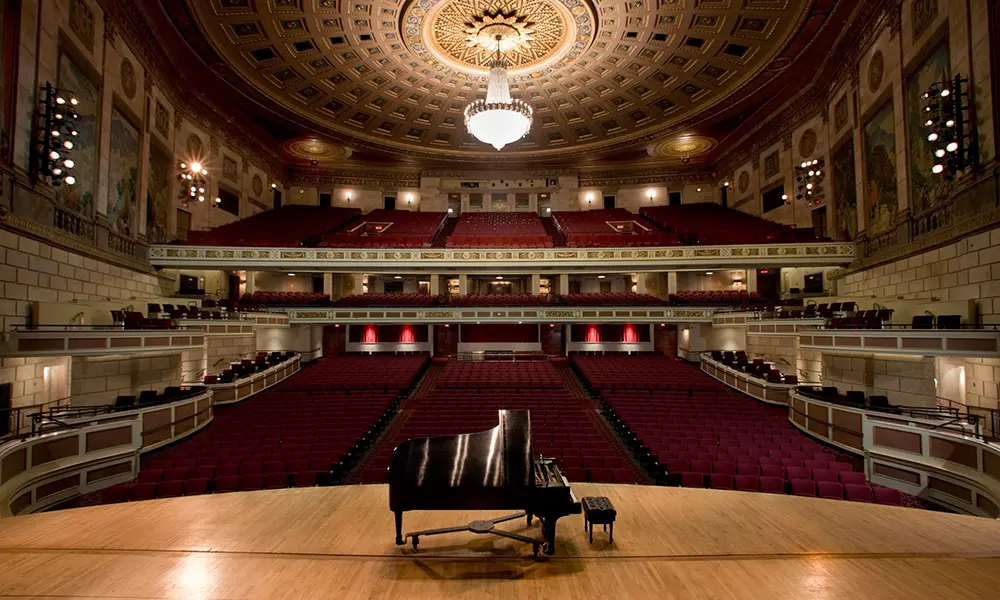 View of a grand concert hall with a black grand piano on stage and rows of red seats.