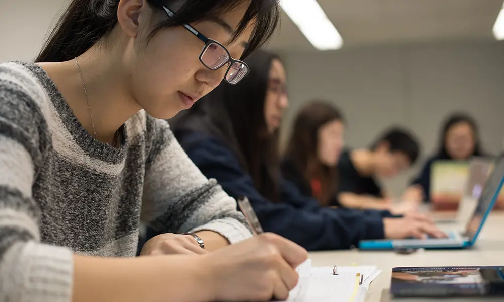 Student in a classroom sits at a table and takes notes in their notebook.